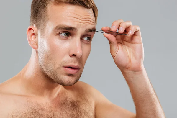 Hansome serious young man removing eyebrow hairs with tweezers