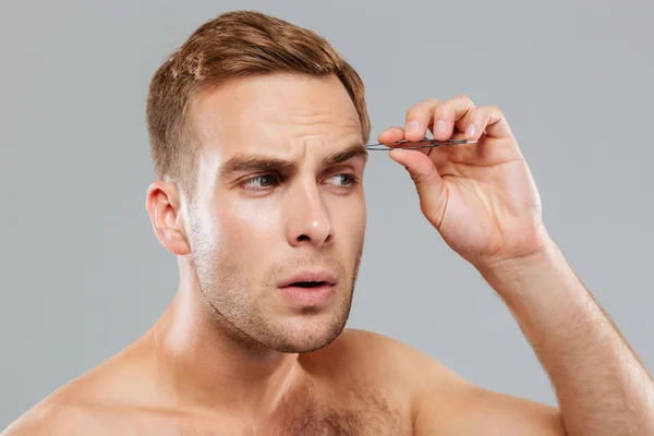 Closeup of concentrated young man removing eyebrow hairs with tweezers
