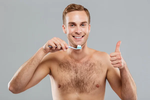 Cheerful young man holding toothbrush and showing thumbs up — Stock Photo, Image