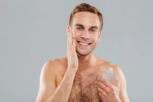 Smiling young man applying lotion after shave on face — Stock Photo, Image