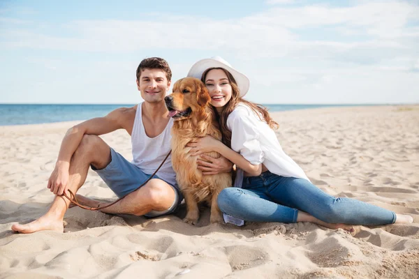 Jovem casal sorridente no amor sentado na praia com o cão — Fotografia de Stock