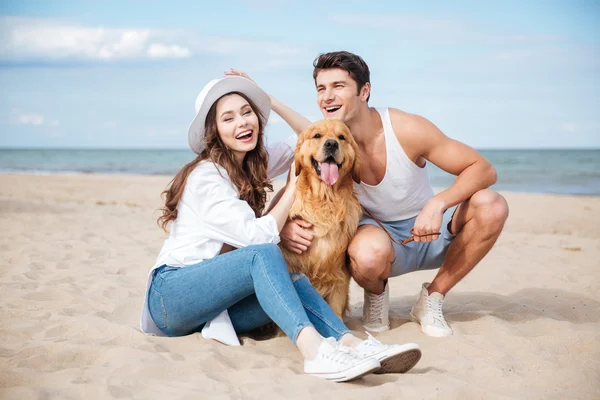 Jovem casal atraente com um cão sentado na praia — Fotografia de Stock