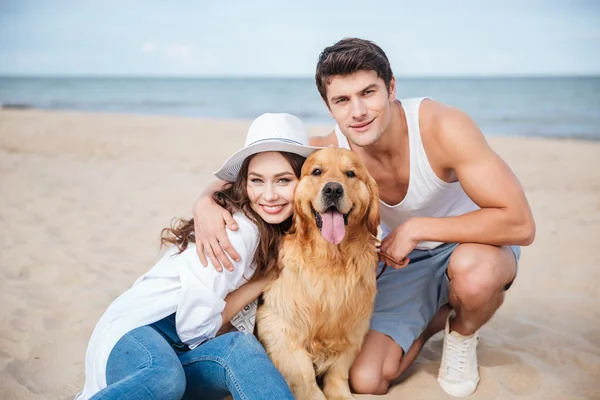 Jovem casal atraente com um cão sentado na praia — Fotografia de Stock