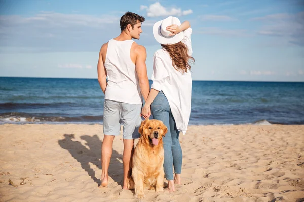 Casal olhando e de mãos dadas perto de seu cão na praia — Fotografia de Stock