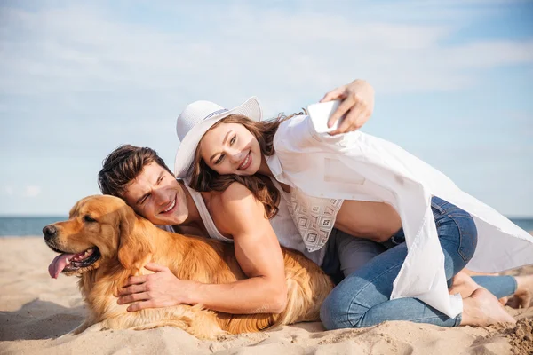 Par med hund pratar selfie med smartphone på stranden — Stockfoto