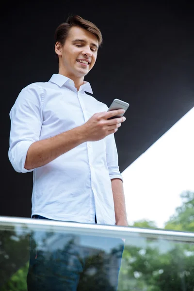 Sonriente joven empresario atractivo de pie y utilizando el teléfono inteligente — Foto de Stock