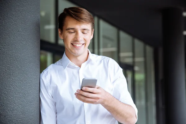 Sonriente joven empresario de pie y utilizando el teléfono celular — Foto de Stock