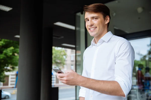 Joven empresario alegre usando el teléfono inteligente y sonriendo cerca del centro de negocios — Foto de Stock