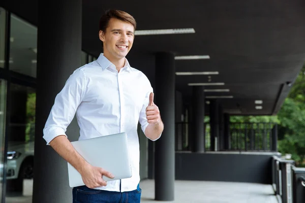 Sonriente hombre de negocios sosteniendo portátil y mostrando los pulgares hacia arriba — Foto de Stock