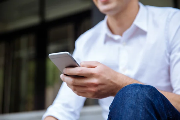 Biznesmen w białej koszuli siedzi i przy użyciu telefonu komórkowego na zewnątrz — Zdjęcie stockowe