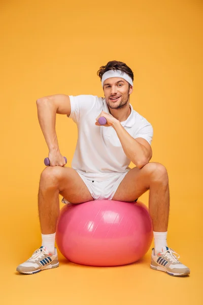 Esportista sorridente sentado na bola de fitness segurando halteres — Fotografia de Stock