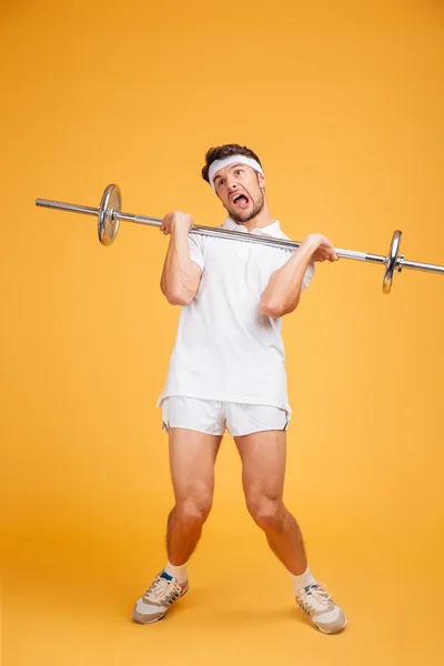 Full length of funny young fitness man holding heavy barbell