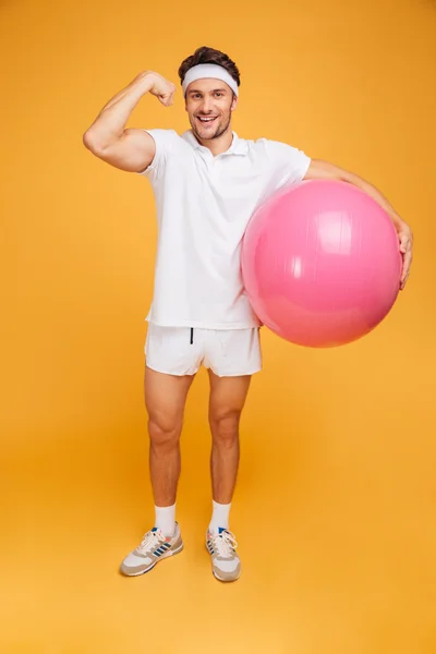 Jovem desportista bonito segurando bola de fitness e mostrando bíceps — Fotografia de Stock