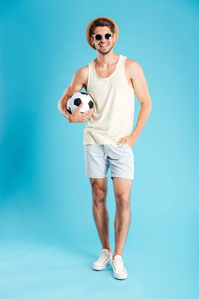 Hombre sonriente en pantalones cortos, sombrero y gafas de sol sosteniendo pelota de fútbol — Foto de Stock