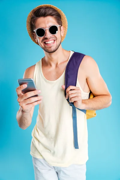 Wesoły człowiek w kapelusz i okulary przeciwsłoneczne z plecaka za pomocą smartfona — Zdjęcie stockowe
