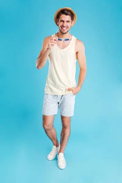 Cheerful young man in hat and shorts holding sunglasses — Stock Photo, Image