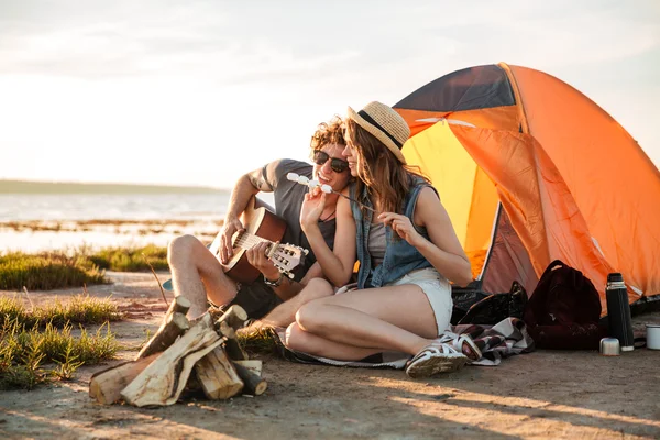 Casal tocando guitarra e comendo marshmallows fritos perto da tenda turística — Fotografia de Stock