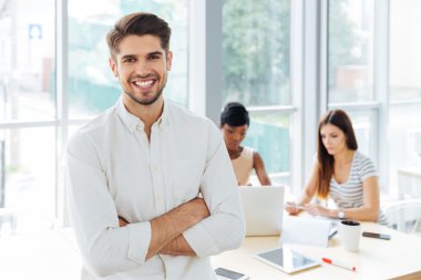 Smiling businessman standing with arms crossed in office clipart