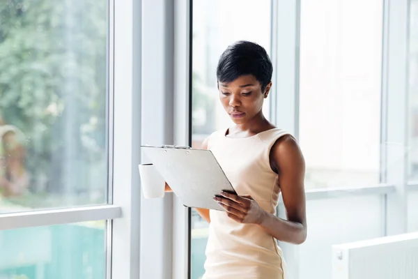 Hermosa mujer de negocios joven africana sosteniendo portapapeles y leyendo en la oficina — Foto de Stock