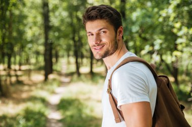 Smiling young man with backpack walking in forest clipart