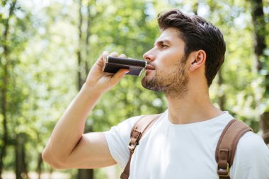 Man with backpack standing and drinking from flask in forest clipart