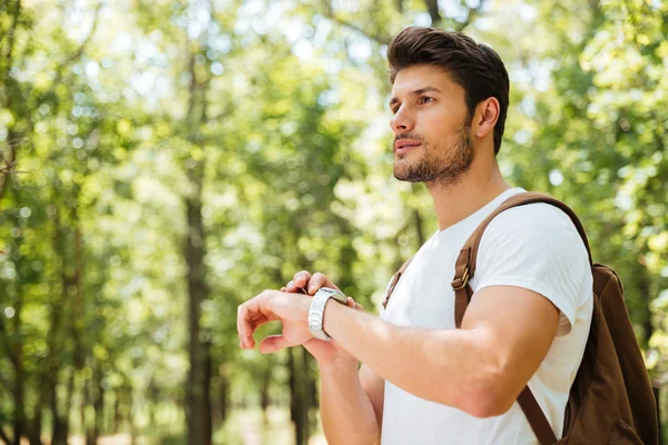 Bystrý mladý muž s batohem, při pohledu na hodinky v lese — Stock fotografie