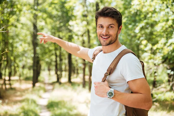 Cheerful young man with backpack pointing away in forest — Stock Photo, Image