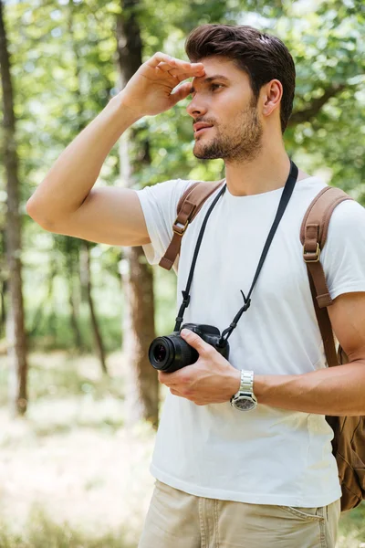 Hombre fotógrafo sosteniendo la cámara y mirando lejos en el bosque —  Fotos de Stock