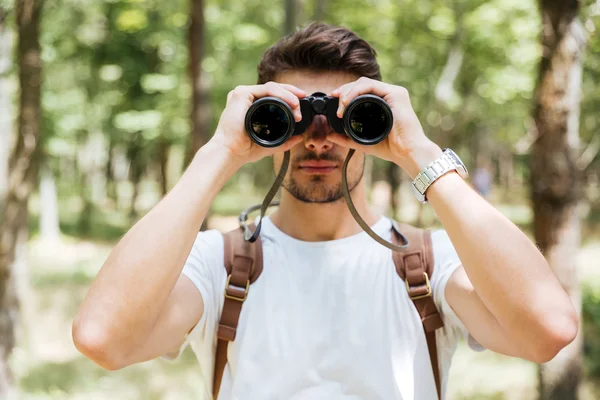 Serious young man with backpack using binoculars in forest — Stock Photo, Image
