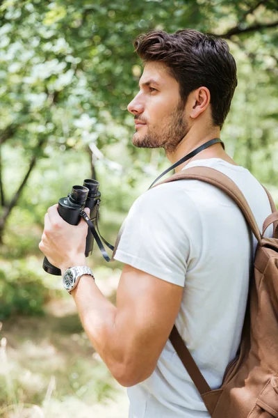 Zamyšlený mladý muž s batohem drží dalekohled v lese — Stock fotografie