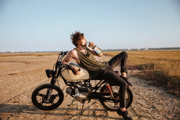 Joven hombre brutal acostado en su motocicleta y agua potable — Foto de Stock