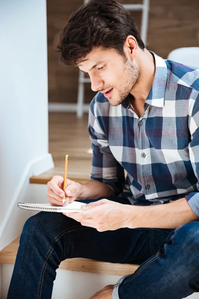 Oturan ve evde Not Defteri'nde yazma genç adam konsantre — Stok fotoğraf