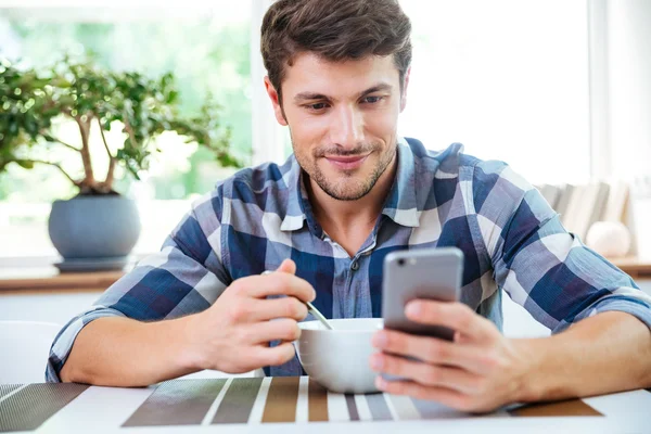 Uomo sorridente che usa il cellulare e fa colazione in cucina — Foto Stock