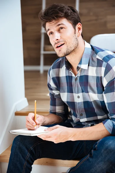 Nachdenklicher junger Mann denkt und schreibt zu Hause in Notizblock — Stockfoto
