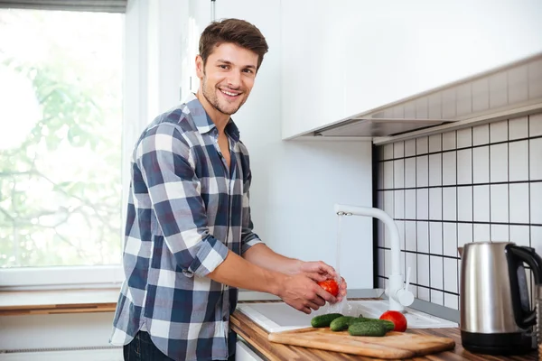 Веселый молодой человек моет овощи на кухне — стоковое фото