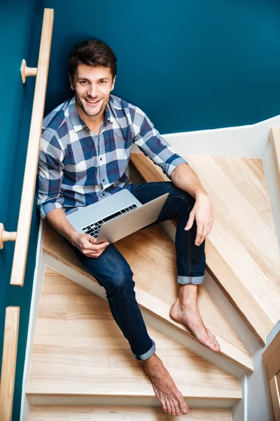 Homem alegre sentado nas escadas em casa e usando laptop — Fotografia de Stock