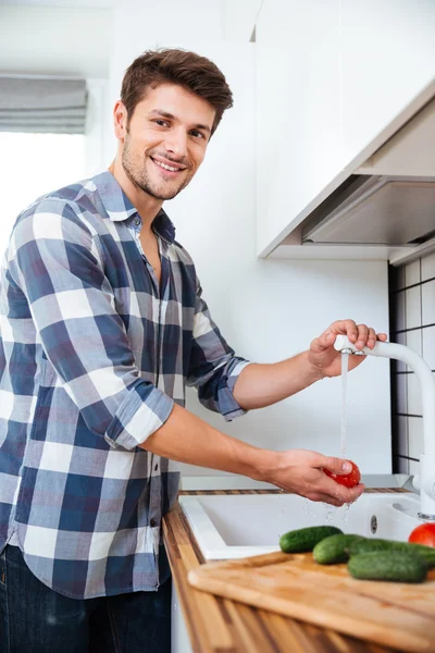 Glad ung man stående och tvätta grönsaker på köket — Stockfoto