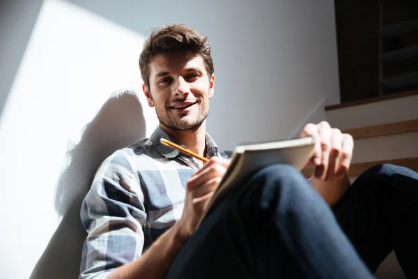 Happy young man sitting the floor and writing in notepad — Stock Photo, Image