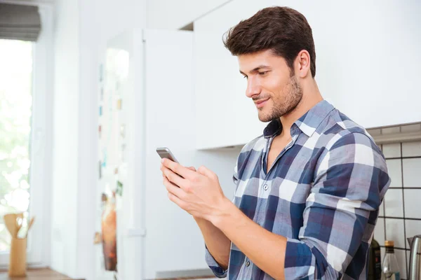 Bello giovane uomo in piedi e utilizzando smartphone in cucina — Foto Stock