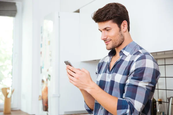 Bonito jovem de pé e usando smartphone na cozinha — Fotografia de Stock