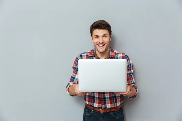 Jovem alegre em xadrez camisa de pé e segurando laptop — Fotografia de Stock