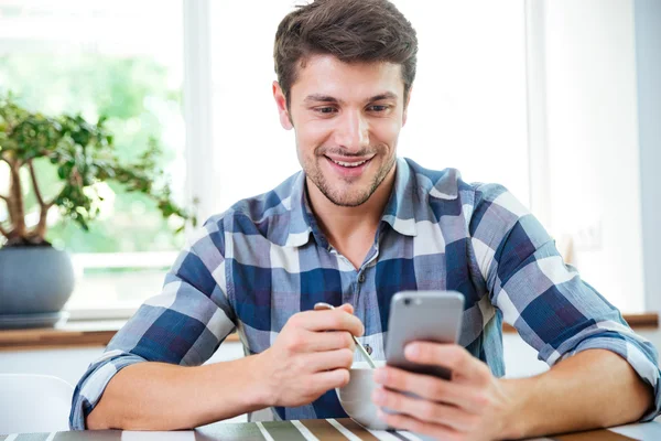 Homem feliz usando smartphone e tomando café da manhã na cozinha — Fotografia de Stock