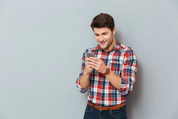 Veselý mladý muž v kostkované košili stojící a pomocí smartphonu — Stock fotografie