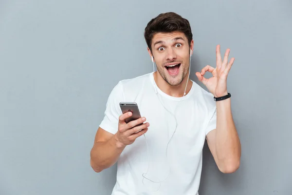 Веселый человек с наушниками, держащий смартфон и показывающий знак "хорошо" — стоковое фото