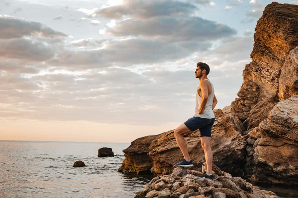 Jovem atleta homem bonito de pé na praia rochosa — Fotografia de Stock