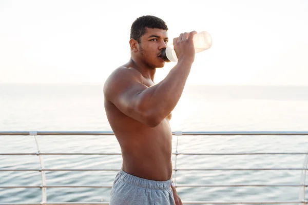Afrikanischer Sportler ohne Hemd steht und trinkt Wasser auf Seebrücke — Stockfoto
