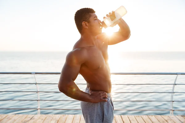 Hemdlos afrikanischer muskulöser junger Mann trinkt Wasser auf Pier — Stockfoto