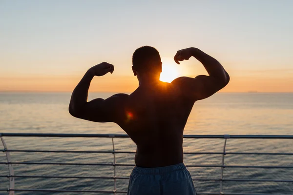 Silueta atletické mladíka ukazuje biceps na sunnrise — Stock fotografie