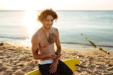 Genç yakışıklı kıvırcık kullanarak sörf tahtası üzerinde otururken smartphone