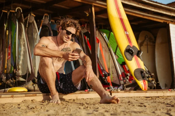 Joven surfista guapo sentado y utilizando el teléfono inteligente al aire libre — Foto de Stock